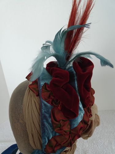 The base is made from a buckram frame that was covered with two-tone velvet in turquoise and rust.  Rust velvet puffs adorn the top, along with rust and turquoise feathers wired into place.