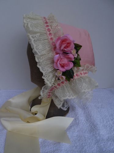 This frothy pink bonnet is exuberantly trimmed with three types of cream lace, and hand tinted flowers on the left.