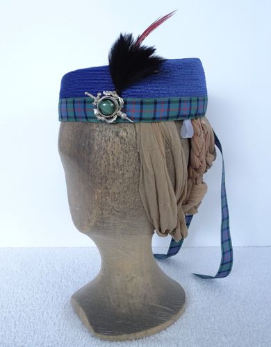 The hat is made of very fine strips of blue straw and trimmed with plaid ribbon, a silver and green brooch, and a feather flourish.