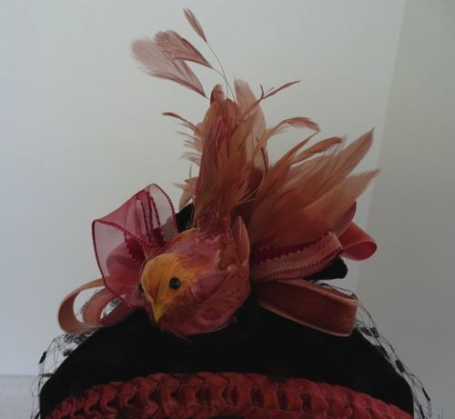 Detailed view of decorations: a hand-tinted bird is placed at centre front, backed by ribbon loops and stripped cocque feathers.